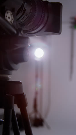 Vertical-Video-Close-Up-Of-Film-Or-Video-Camera-On-Tripod-Shooting-Movie-In-Studio-With-Light-In-Background